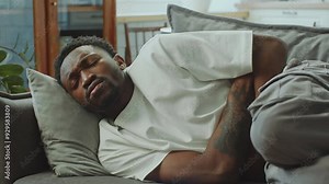 Young sick Black man lying on couch in fetal position as suffering from stomach ache or nausea during the day at home