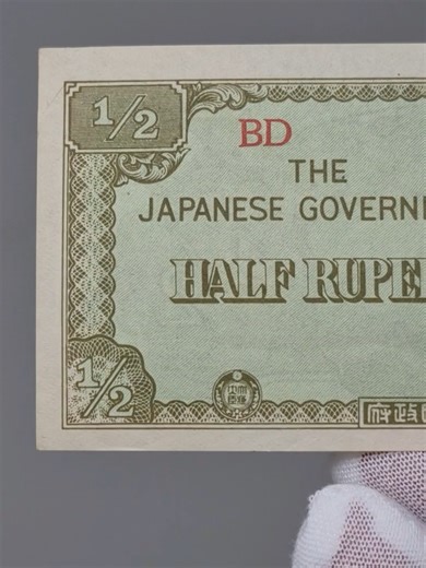 1942 Japan Half Rupee Burma Banknote Japanese Invasion Money Obverse: Issued by The Japanese Government for circulation in occupied Burma during World War II. The design features a view of Burmese temples and palm trees at right, with denomination “½” in all four corners. The text reads “THE JAPANESE GOVERNMENT – HALF RUPEE.” Series code “BD” printed in red. Decorative border with Japanese characters at bottom center. Reverse: Ornamental guilloche patterns with denomination “½” prominently displ
