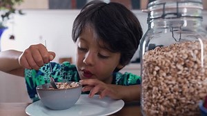 Boy enjoying a spoonful of cereal at the dining table, mouth wide open. Jar of cereal in the foreground emphasizes the wholesome and nourishing breakfast moment