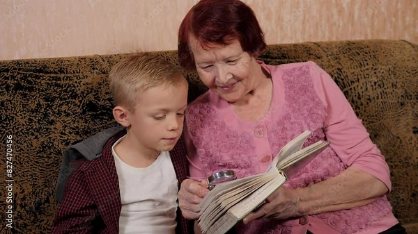 Close-up of an elderly grandmother reading a book with her grandson using a magnifying glass while sitting at home on the couch.