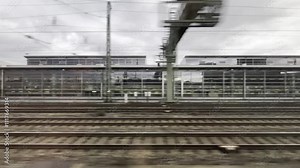 View from the window of a train passing a large station. Various buildings and railroad tracks fly by. From the train's window you can see the city and oncoming and passing trains.