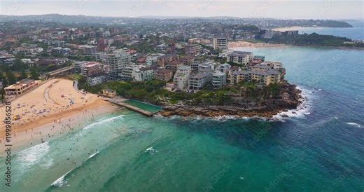 Dee Why beach, popular tourist destination with resort hotel buildings, bustling sand shoreline, people swimming and relaxing, prominent rock pool, vibrant Sydney coastline. Aerial view drone panorama