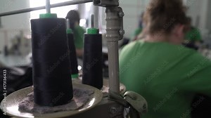 Sewing machine pulling black threads of spools in focus, group of female dressmakers stitching clothes in workshop inthe blurred background, steadicam shot, close up, room interior,
