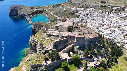 Aerial drone video of iconic archaeological site of Acropolis of Lindos built uphill featuring picturesque village, amazing organized beach and Saint Paul bay, Rhodes island, Dodecanese, Greece