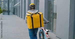 Rear of African American male courier with big bag over back in casque walking and leading bicycle on street and delivering food or parcels. Back view on deliveryman bicyclist have a walk with bike.