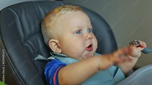 A little boy is sitting in a high chair, eating porridge, and his mother is trying to feed him with a spoon. Baby food. The first meal for a child.