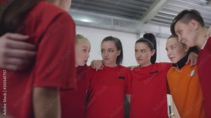 Team of young female soccer athletes in sports uniform standing in circle, embracing and listening to leader while getting ready for match in locker room