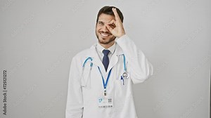 Cheerful young hispanic doctor having fun, flashing a joyful smile while doing an ok sign with fingers. peeping through his hand, isolated against a stark white background.