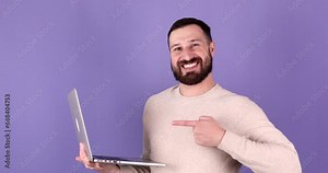 Young happy bearded caucasian IT man hold in hands typing use work point index finger on laptop pc computer isolated on purple background. People lifestyle concept.