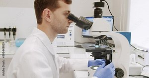 Male doctor using microscope in laboratory. Side view of man in lab coat and latex gloves using microscope to examine sample during work in clinic in daytime.