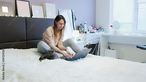 young woman is sitting on the bed in the bedroom with her cat, looking at the laptop screen, typing, or chatting online with friends. Attractive brunette girl at home uses a computer, self-isolation,