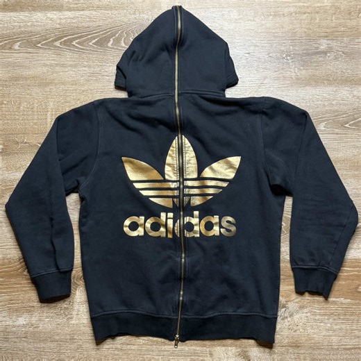 2015 Jeremy Scott x adidas black & gold full back zip-up hoodie. Features the Jeremy Scott logo on the front chest and the adidas trefoil logo on the back, both in a metallic gold foil. The standout feature of this piece is a functional two way zipper running down the center of the back and on the hood. This detail was added as an aesthetic choice where it utilized more fashion rather than practicality. You can half zip the hood or unzip it all the way depending on your mood #jeremyscott #adidas