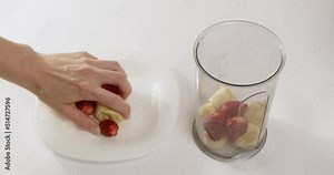 Hand of woman takes fresh fruits from plate throwing into plastic container on table. Housewife prepares dessert with banana and strawberry closeup