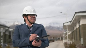 Serious urban development inspector in a suit and tie conducting an audit of renovation work on suburban street making notes on report on digital tablet with stylus