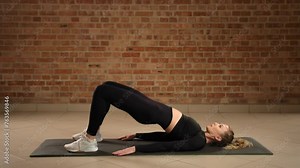 Performing hip bridges on a mat, the dedicated trainer is targeting glute muscles and lower back, promoting better posture and core stability in a serene gym setting with a brick wall background.