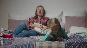 Charming mother watching tv, changing tv channels with remote control and cute preadolescent daughter watching video content online on mobile phone while family relaxing on bed in the evening.