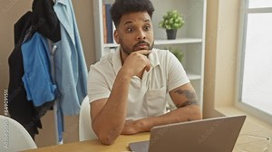Thoughtful man with tattoo using laptop at home office, reflecting diversity and modern work environment.
