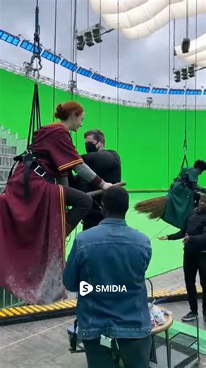 Behind The Studio on Instagram: "Harry Potter BTS: Flying Lessons in the Sky ✨🎬 #shorts #harrypotter #bts Actors suited in Hogwarts Quidditch robes are strapped into harness rigs as the crew prepares them for high-flying broomstick shots. A massive green screen curves around the entire set, ready to transform into a sweeping view of the Hogwarts grounds. Costume artists adjust capes, while technicians secure safety wires and position the broom props under each actor. Studio lights beam from abo