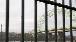 Runcorn silver jubilee bridge parallax behind passing iron park railings & waterfront promenade tree open space.