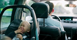 Closeup crying baby boy sitting in the baby car seat on the rear seat inside of car, while car is riding along the road. Father behind him is driving. Slow motion