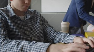 Medium close-up tilt shot of geeky 12-year-old Caucasian male school student in checked shirt sitting in programming class, writing computer code on laptop, male teacher assisting girl in background