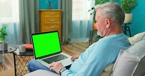 Older Man at Home Sitting on a Couch Works on a Laptop Computer with Green Mock-up Screen. Man Using Computer, Browsing through Internet, Watching Content, Chatting in Social Networks with Friends.