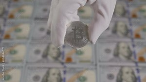 Close-up of a hand in white gloves holds a silver bitcoin coin against the background of one hundred dollar bills. Digital money, banking, investment, finance and business concept.