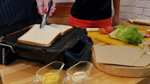 Cooking a sandwich in the kitchen. Hands with tongs take white toast bread from a wooden stand and place it to be fried in a sandwich maker