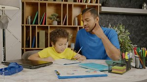 Caring attractive African American father checking and explaining school assignment, helping smart cute school age son to do homework while family studying together in domestic room,