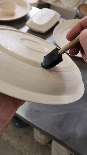 Before glazing, I coat the bottoms of my pots with Forbes wax using a 1" foam brush, in this case on a new chip-and-dip platter. The wax creates a resist that keeps glaze from adhering to the clay surface when glazed. A damp sponge removes any glaze residue that remains after the piece is dipped. This is called 'dry footing'. Without it, the glaze on the piece would fuse to the kiln shelf during firing—ruining both the piece and the shelf. It's a simple step, but an essential one. | Hamilton Wil