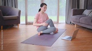 Beautiful woman sit and do yoga posture or move by torsion on side and she look example from laptop in home living room. Concept of workout at home for good health and prevent covid virus infection.