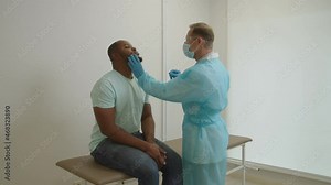 Middle aged male physician wearing PPE holding COVID-19 swab collection stick, taking nasal and oral specimen of african american man for PCR test for coronavirus at medical clinic.
