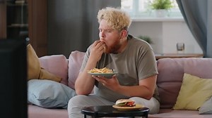 Young obese man sitting on sofa at home, eating fries with ketchup and watching TV