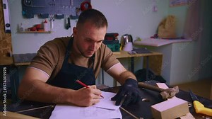 Craftsman wearing black gloves and apron meticulously sketching furniture design on paper at workbench in his workshop, surrounded by tools and materials