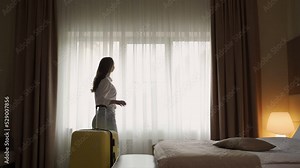 Female tourist pulls back white curtains to enjoy view from window. Young woman stands near suitcase in hotel room backside view slow motion