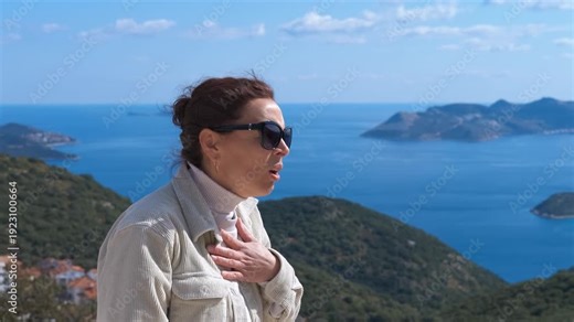 Woman with shortness of breath using asthma inhaler outdoors. Mature woman experiencing breathing difficulties and an asthma attack, using a preventer inhaler while standing on a mountain with a sea