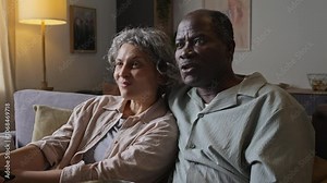 Tilt shot of happy diverse middle-aged married couple enjoying watching TV together at home, husband hugging wife from behind, spending leisure time together