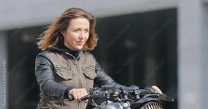 Woman sits on motorcycle posing vintage motorbike stroking hair sunny urban defocused background. Female biker tries to smile eyes welling up of dazzling sun holding handlebars touch red hair close up