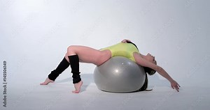 Beautiful girl in a yellow sports suit and black leggings is engaged in gymnastics on a gray rubber ball, swimsuit, beautiful skin, figure, barefoot, crouchs on the ball, jumps, slog