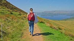 Following a hiker walking on a mountainside trail surrounded by grassy fields and majestic views, Conic Hill, Scotland
