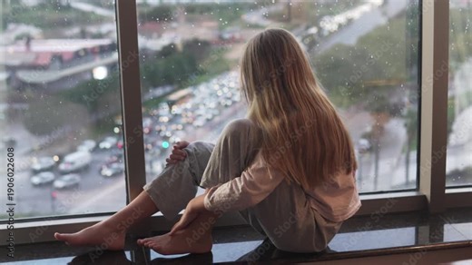 Young Girl in pajamas sitting barefoot on a windowsill and looking at the city through a rainy window