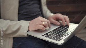 Man is using touchpad and keypad of notebook, close-up of hands. He is holding it on laps and sitting outdoors