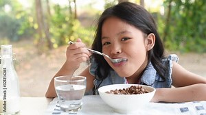 Cute little asian girl with milk and healthy breakfast at table in kitchen. enjoying healthy breakfast eating cereal and drinking milk at bright sunny kitchen next to big garden view window