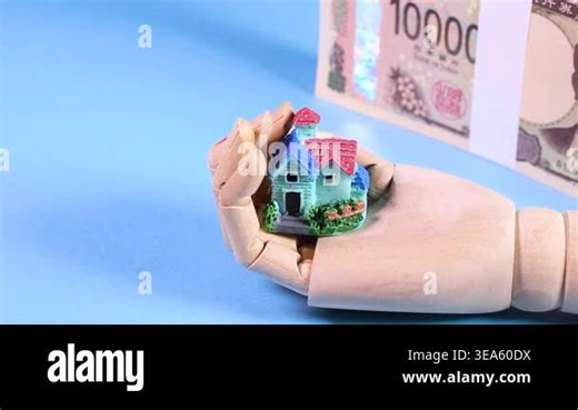 Rotating stack of 10,000 yen Japanese banknotes and a miniature house on a wooden hand against a light blue background, with text "Bank of Japan" and "Bank of Japan Note Stock Video Footage - Alamy