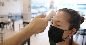 close up shot of staff using infrared thermometer checker on young Asian woman wearing facial protective mask customer's forehead before enter, waiting for Temperature checkpoints in restaurant,