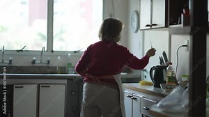 Elderly Woman in Home Kitchen Retrieves Cooking Utensils from Drawer and Knife Holder, Heading towards Sink for Meal Preparation