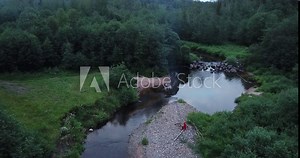girl in a red cloak sits by a fire on the bank of a river with a waterfall. A fire burns, smoke rises. Fabulous image, fairytale character.