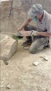 A stonemason is shaping a rough block of stone with an angle grinder creating significant dust