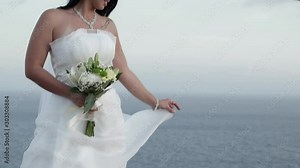 Newly wedded bride posing on top of rocks and holding wedding dress with flower bouquet on wedding day at sea coast oceanview during bridal photoshoot celebrating love slow motion reveal tilt shot