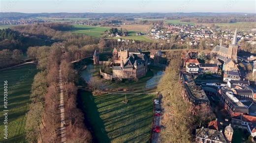 A medieval castle with a wide moat stands beside a tall steepled church, surrounded by clustered homes, open fields, tree lined roads, and forest, forming a harmonious historic landscape.